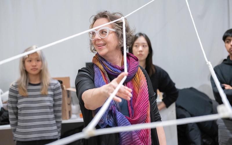Artist Janet Echelman, wearing large white glasses and a colorful pleated scarf gestures while engaging with students during a class activity involving ropes strung in geometric patterns. Several students look on in the background.
